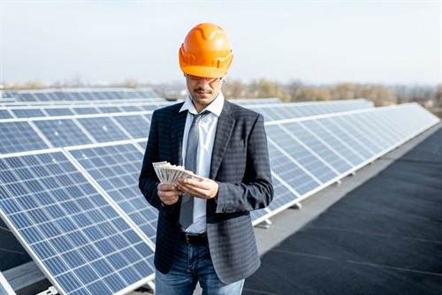 Man with safety helmet holding money on the roof with solar panels